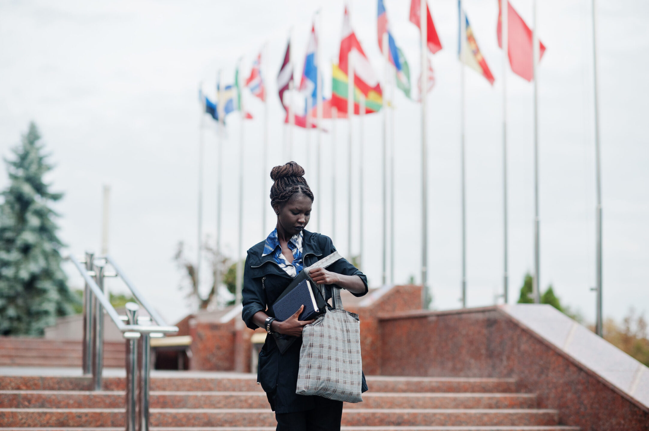 African student female posed with backpack and school items on yard of university, against flags of different countries.