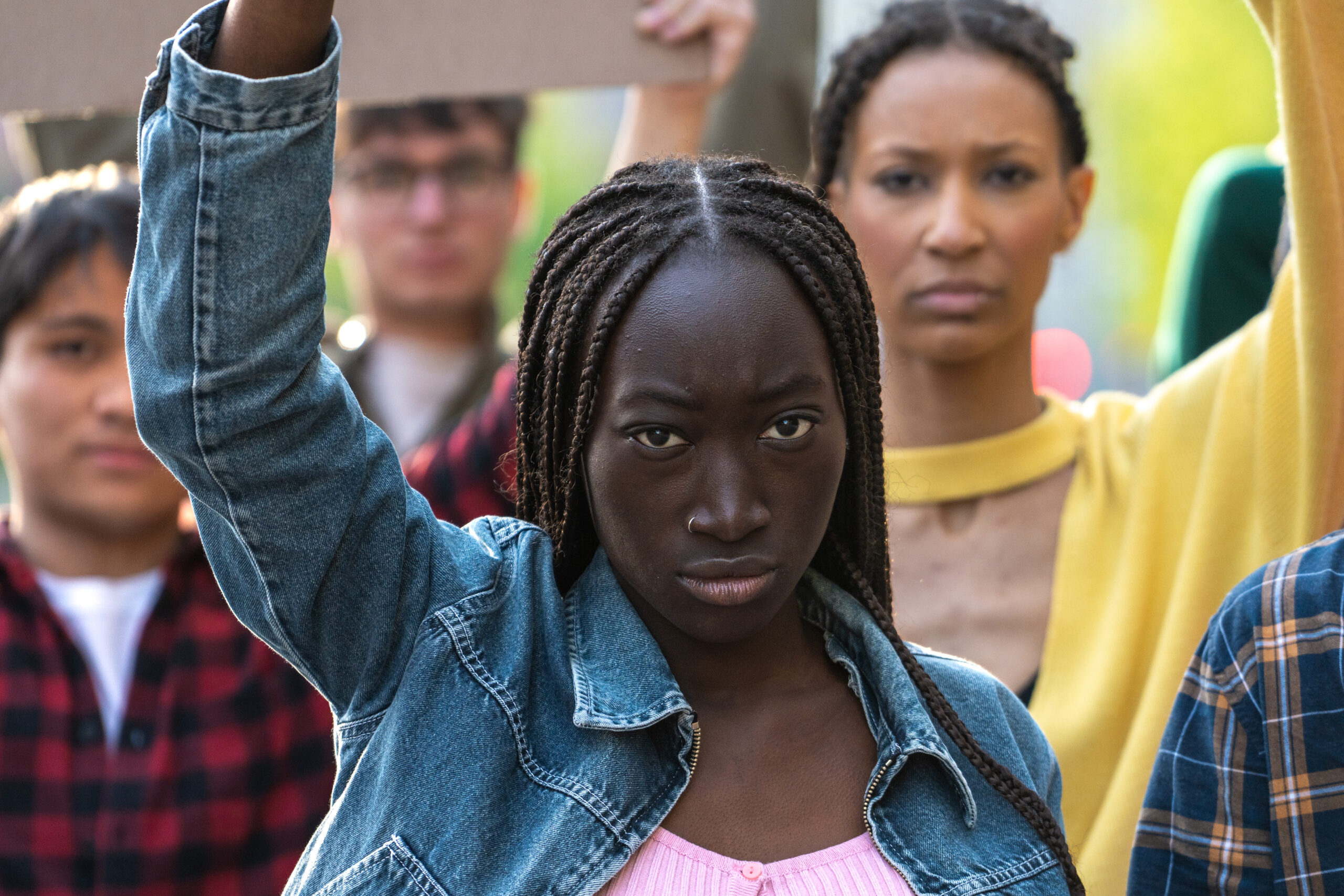 Close-up of a young African woman with raised fist, leading a diverse group of protesters. Image captures strength, determination, and unity in youth activism for social change