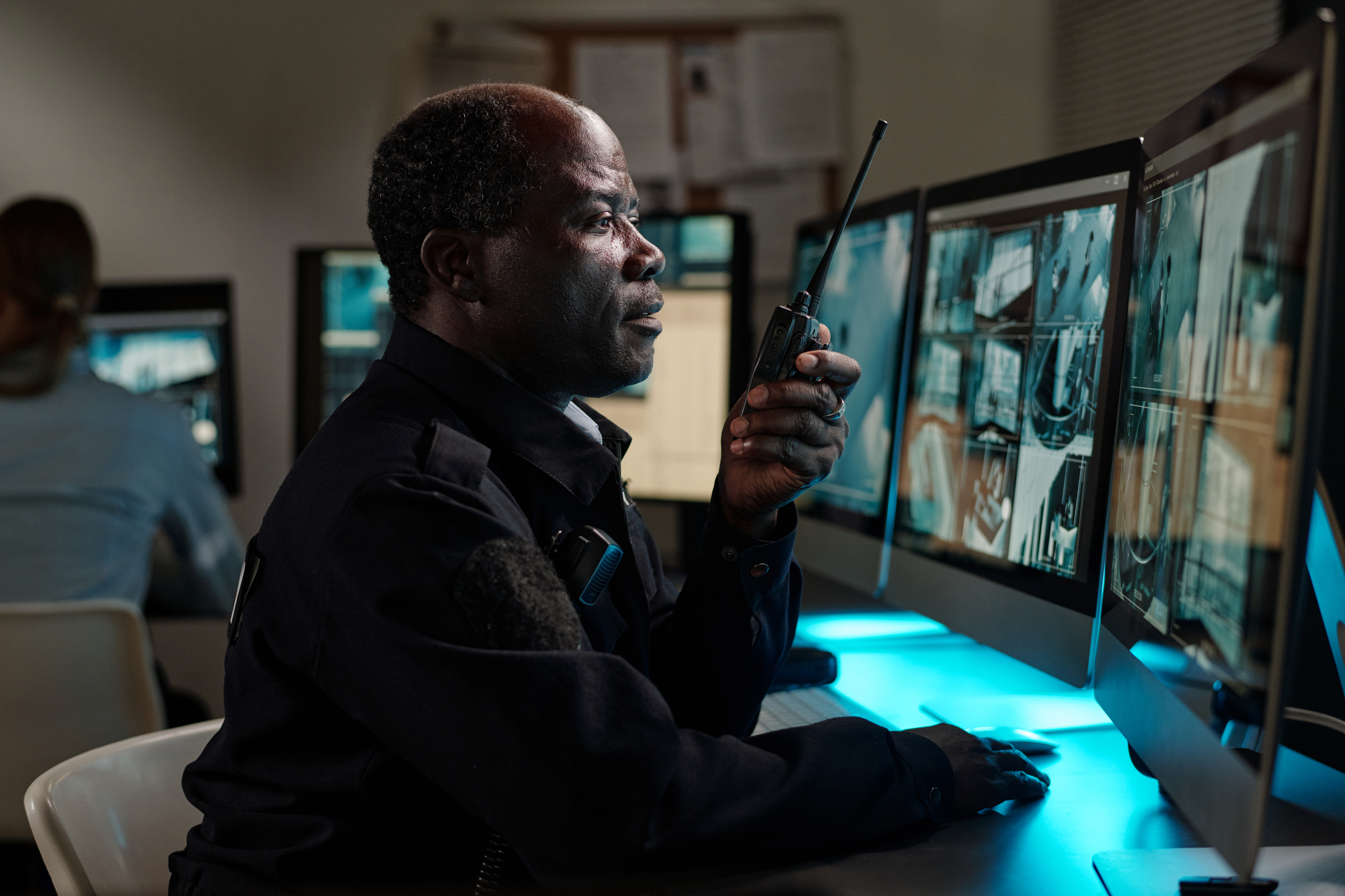 Mature African American male officer sitting in front of computer monitor and looking at screen while speaking on walkie-talkie