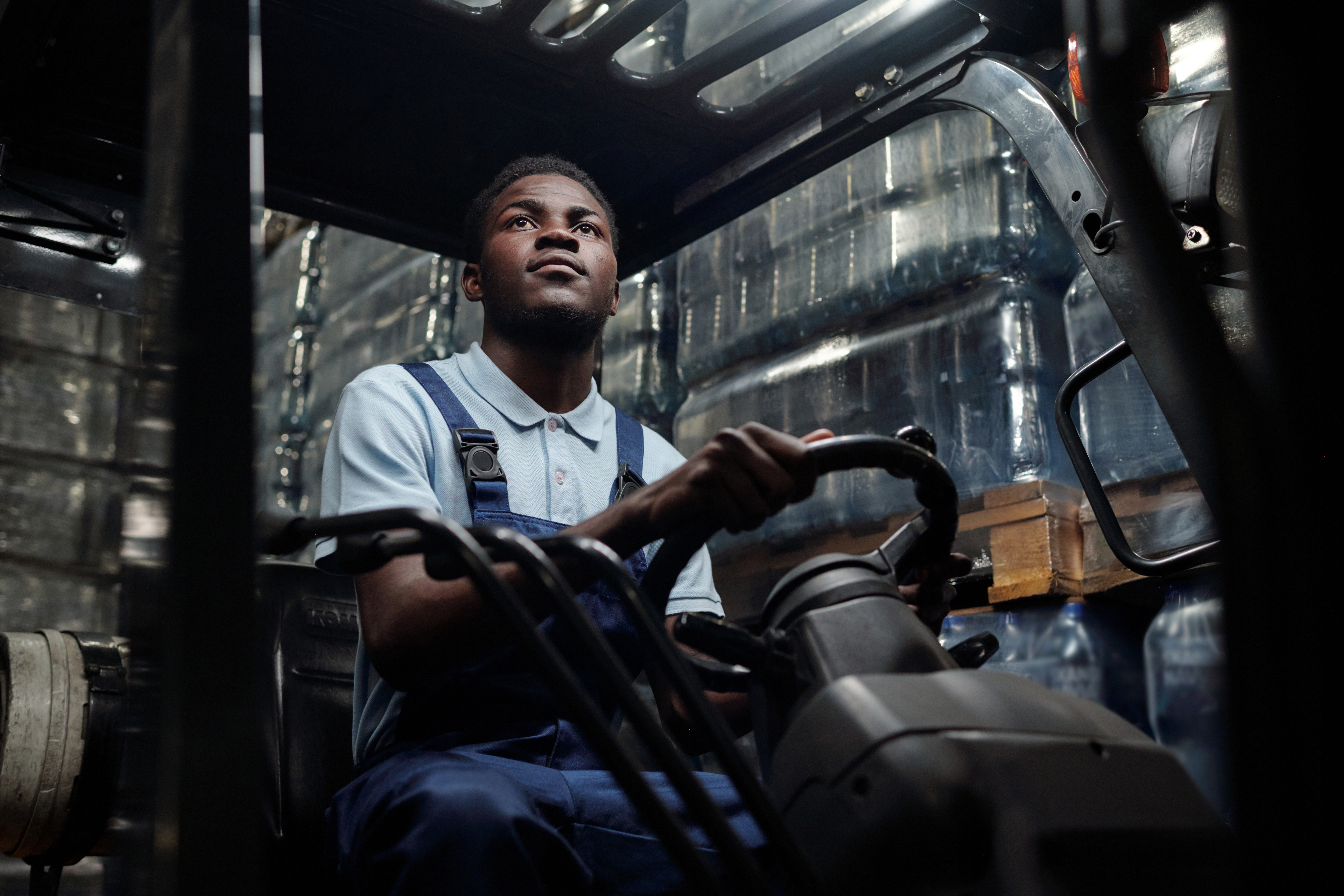 Young worker of huge industrial warehouse in coveralls sitting in forklift and driving it while moving along aisle between stacks of goods